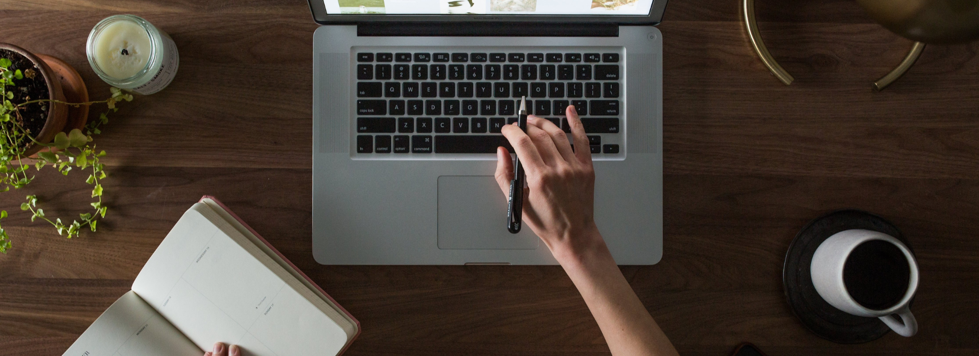 Person typing on keyboard with a coffee cup, notebook, plant, and candle next to them.