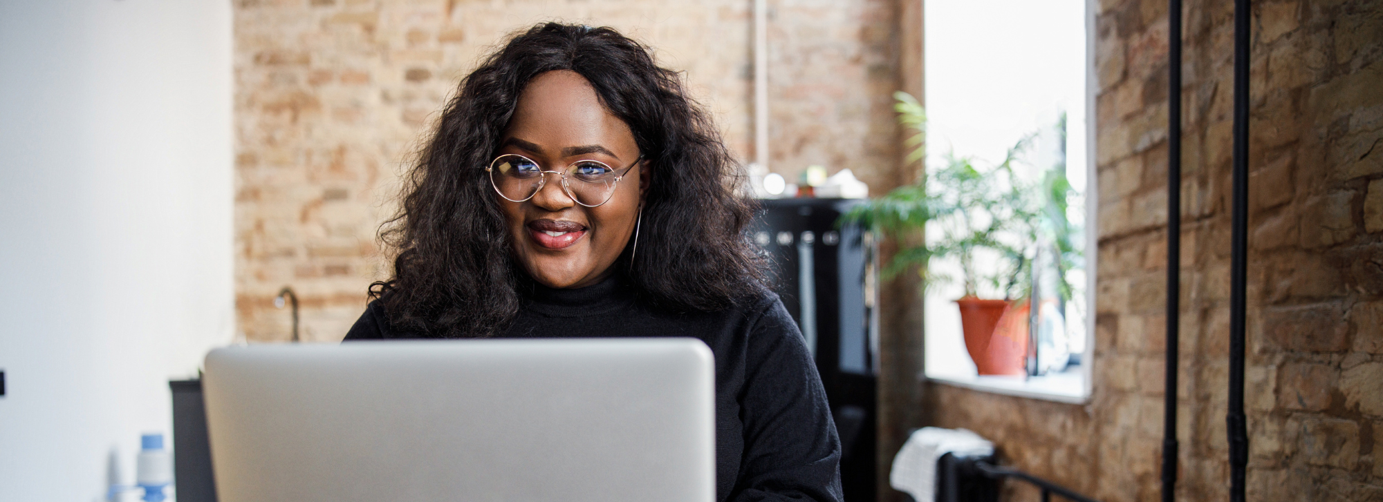 A woman on her computer smiling at the camera. There is a brick wall and a plant in the background.