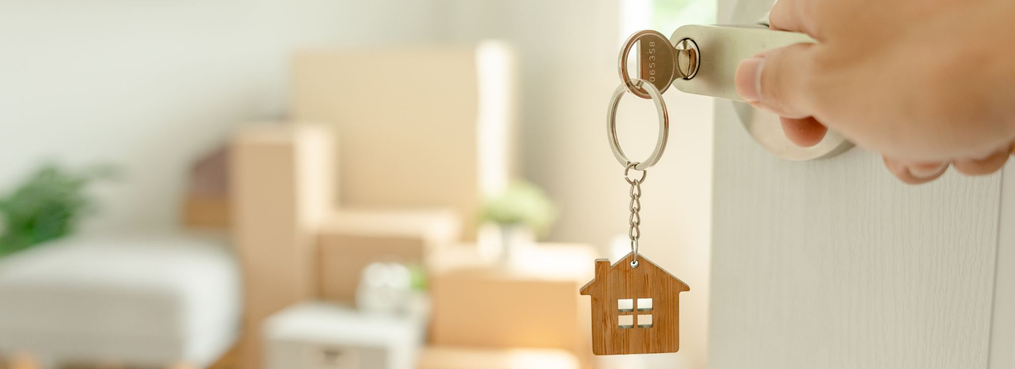 Someone's hand is opening a door that has a key in it. The key has a house trinket hanging off of it, and there are blurred boxes in the background.