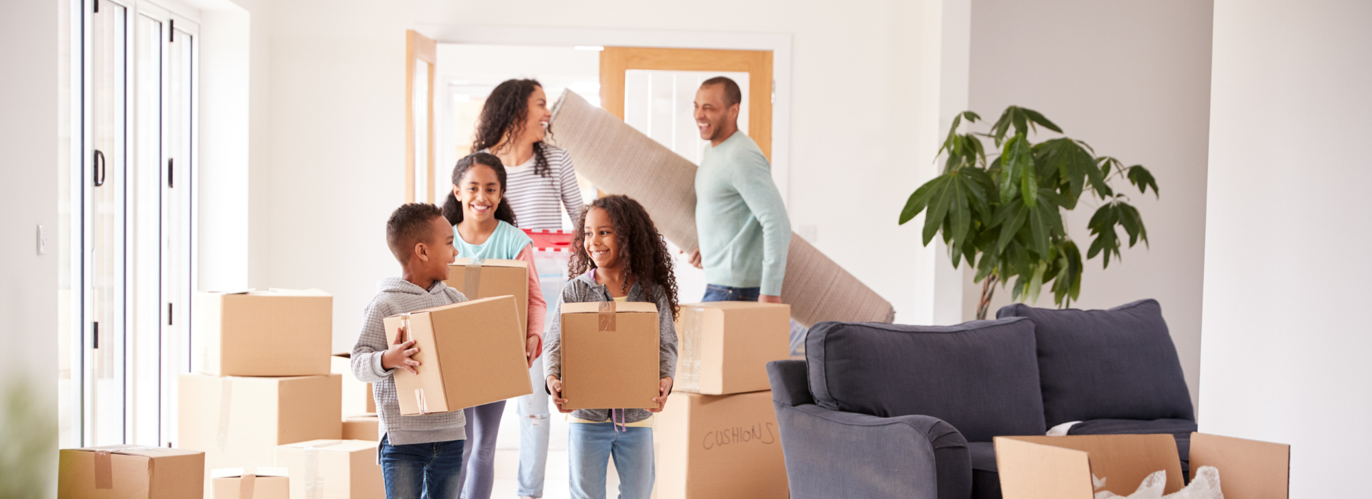 A family of five carrying boxes around the living room of their new home, while smiling.