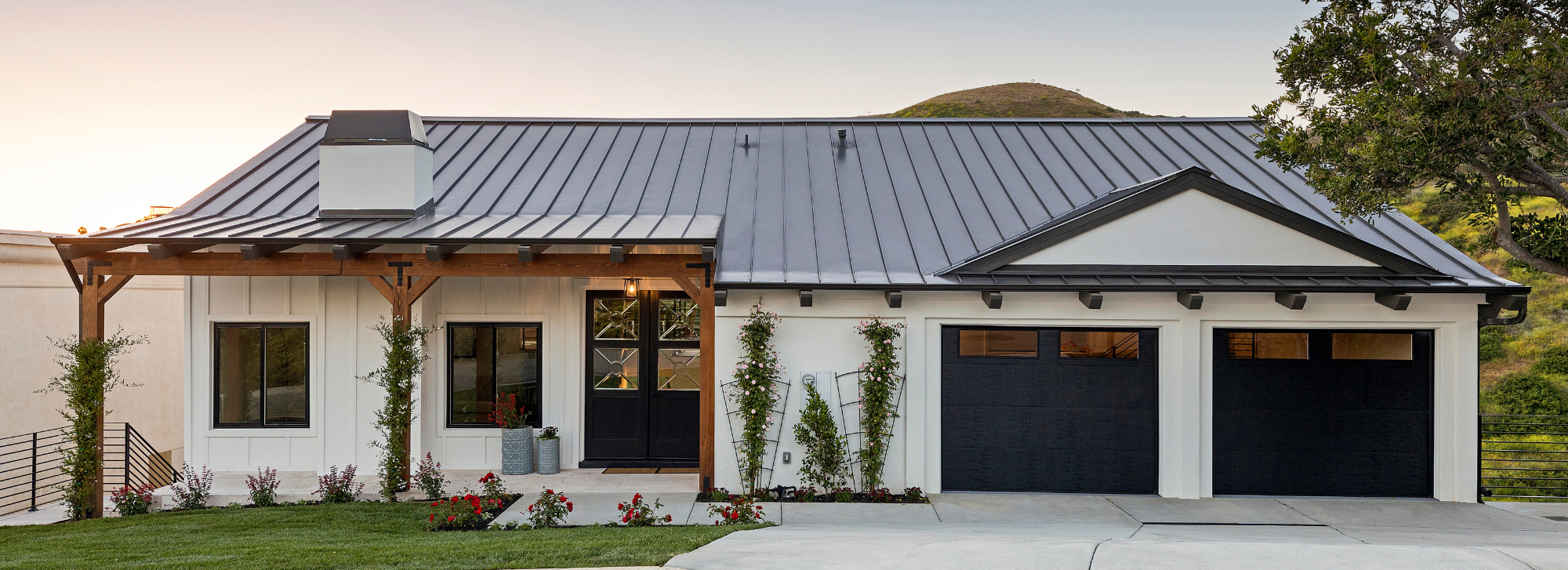 A medium size house that has white siding and a black roof. There are a couple different plants in the front yard.