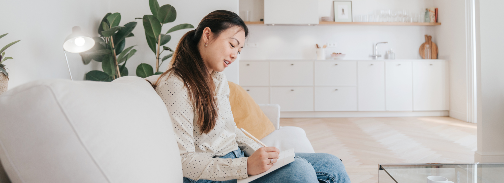 Girl sitting in her living room writing in a journal, and her kitchen is in the background.